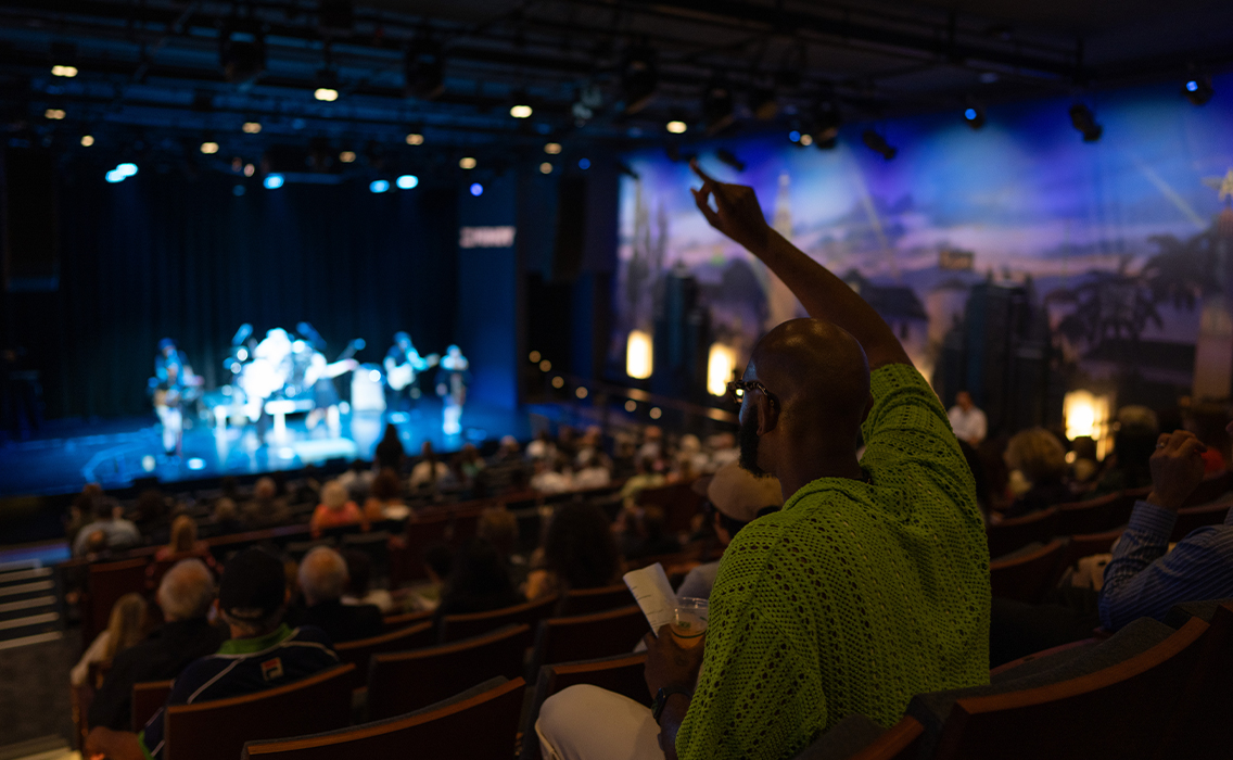 An audience member in a lime green shirt raises their arm in the air while a performance occurs on The Nimoy stage