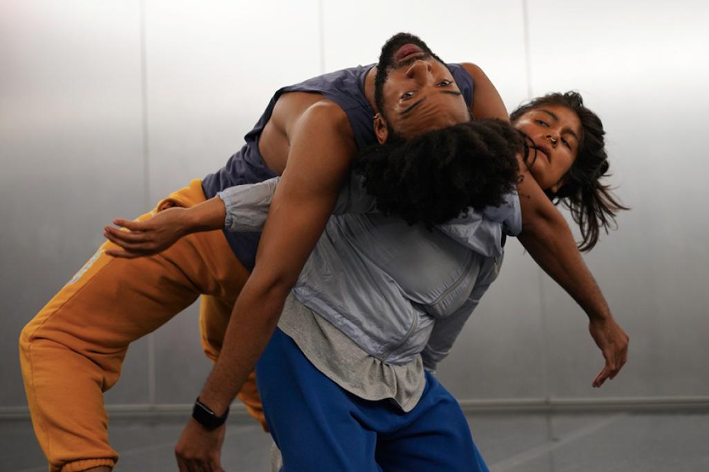 Two dancers pose in front of a grey wall with one leaning backwards on the other performer, who is leaning forwards