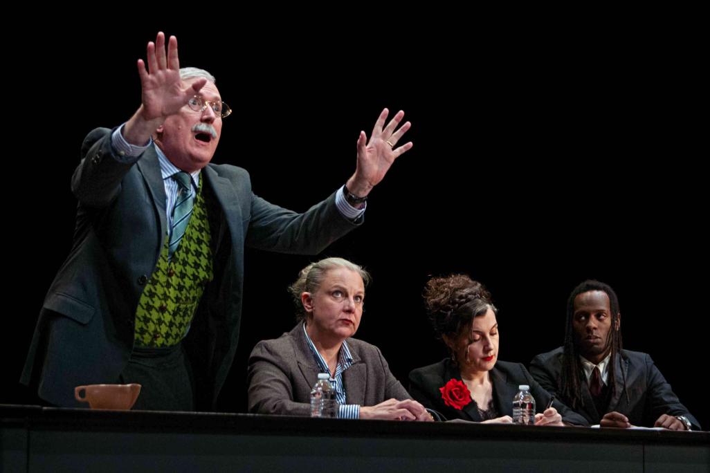 Performers from 'Ulysses' sit at a long table on stage with papers falling from above them