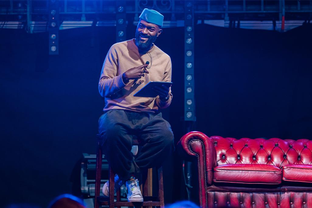 Inua Ellams sits on a stool on stage and speaks into a microphone