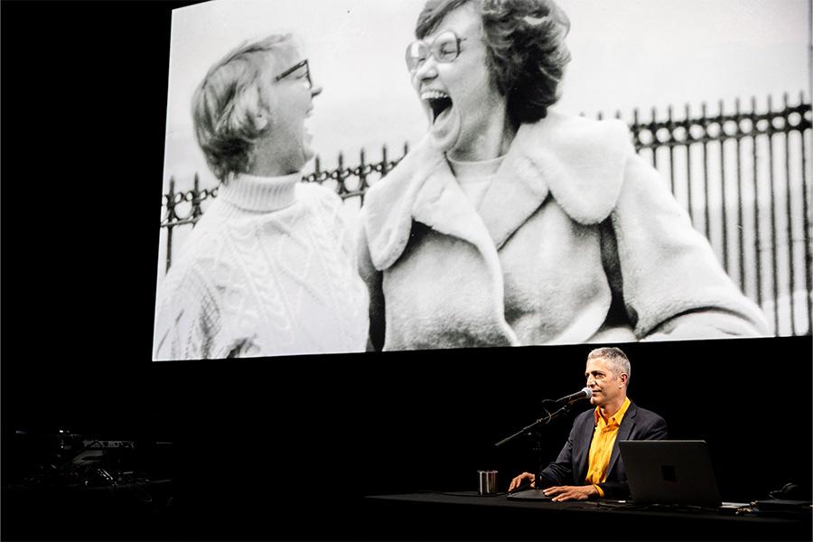 Sam Green on stage with a black and white photo displayed behind him 900x600